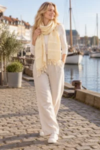 Model går på en havnepromenade med et lyst, gul silkehalstørklæde, hvid bluse og lyse bukser.