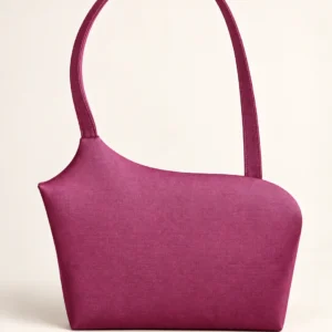 En magenta skuldertaske med asymmetrisk form og lang rem mod lys baggrund.
