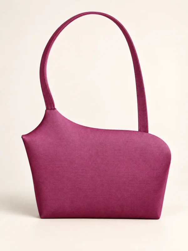 En magenta skuldertaske med asymmetrisk form og lang rem mod lys baggrund.